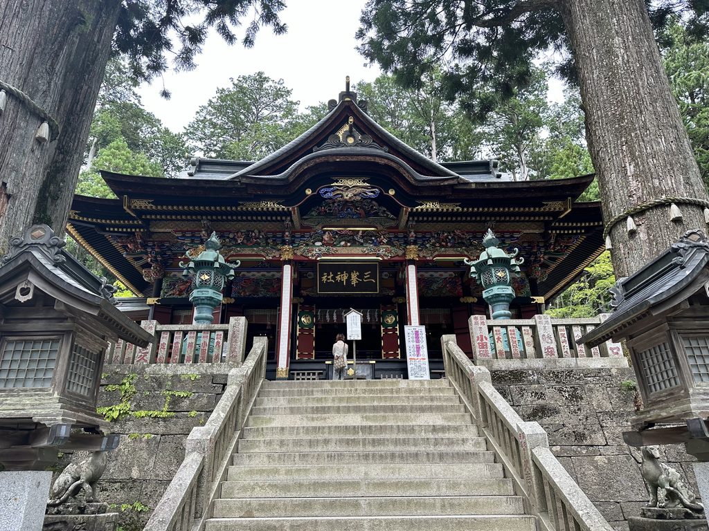 三峯神社の拝殿を石段の下から見上げた様子。極彩色の彫刻と「三峯神社」の扁額、両脇には杉の巨木と狼(お犬様)の石像が見える